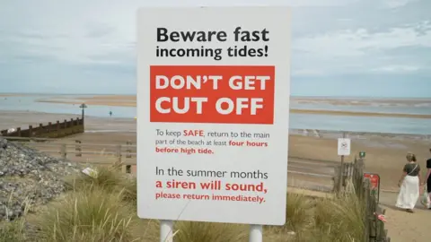 Qays Najm/BBC A red and white sign warning people of fast incoming tides at the beach. It tells people to be vigilant and to listen out for a siren in moments of danger. Behind the sign is the beach, which has water cutting through the sand, creating dunes.