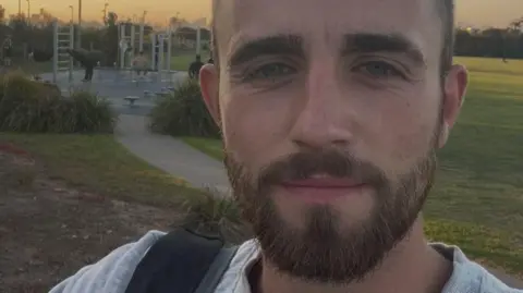 Tom Cowan Tom Cowan smiles down the lens while taking a selfie. He has blue eyes and dark brown hair. Behind him is an outdoor exercise park. 