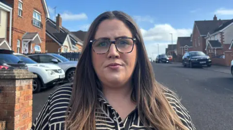 A woman with long, brown hair, wearing black-rimmed, clear glasses stairs into the camera. She wears a black and white striped shirt as she stands in a housing development, with houses either side of her. A variety of cars are parked in drive ways and partially on the footpath outside the houses.