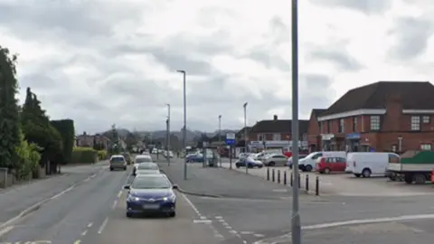 A street with cars driving along and parked beside a row of shops and buildings, with a car park under a cloudy sky.
