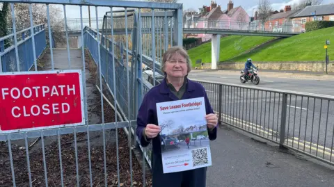 LDRS Campaigner Debbie Hills next to the closed footpath with Leazes Footbridge in the distance. Ms Hills wears a purple coat and glasses and holds a Save Leazes Footbridge poster. A motorcyclist is travelling on the road.