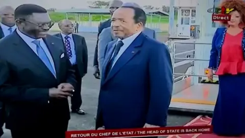 CRTV A screengrab from live television showing Paul Biya being greeted by an official at the airport. His wife, Chantal, can be seen in the background.