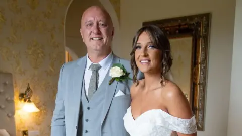A woman with tied up brown hair in a white wedding dress is smiling at a man who is also smiling and wearing a white shiret with a grey vest and tie.