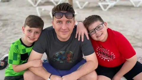 Tim Sadler Image of two boys and a man sitting together cross-legged on a beach. One boy is wearing a green t-shirt, the man is wearing a black t-shirt and blue shorts and sunglasses on his head. The second boy is wearing a red t-shirt and is wearing glasses. All three are smiling at the camera.