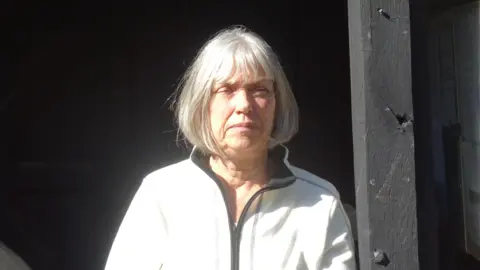 Jacquie Eykelbosch, leaning against a black timber upright. She has bobbed grey hair and is wearing a white top with a black zip. She is squinting slightly in the sun. 