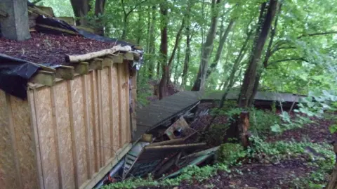 Wiltshire Council A wooden structure made of pale wood sits in the middle of a woodland surrounded by other buildings and greenery