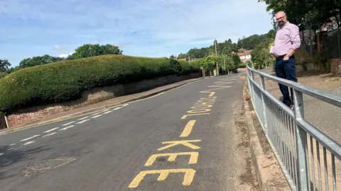 Owen Sennitt/BBC A man stands next to yellow 'school, keep clear' road markings on a sunny day, looking down the road