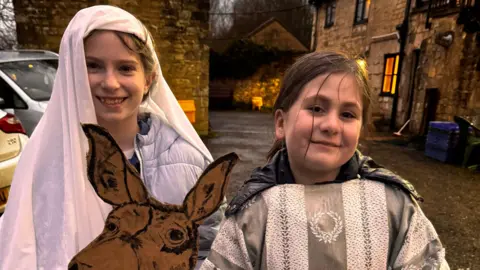 Two children, dressed as Mary and Joseph, with Mary holding a cardboard cutout of a donkey, stood outside of a Cotswold stone house on a late December afternoon, in the damp. Cars can be seen on the driveway behind them, and there are some lights on in the house.