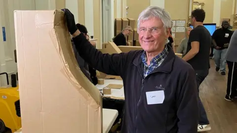 Les Fawcett stands with a curved structure that is part of the arched window he has made. He has white hair and is is wearing a shirt and dark fleece and glasses and is smiling at the camera.
