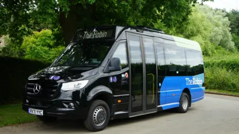 Gloucestershire County Council A black and blue minibus which says 'The Robin' on the side of it. It is parked on the side of a road, next to trees and hedgerows.