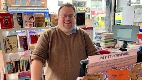 Nik Lowe, who has brown hair, glasses, a goatee beard and is wearing a brown jumper with the sleeves rolled up, smiles at the camera from behind a counter in a shop stacked with books. 