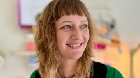 Karina Patfield A woman with long hair smiles and looks to the right. She is wearing a black top and a green cardigan.