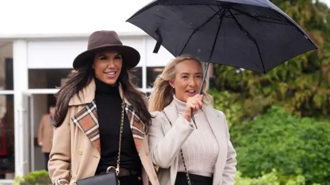 PA Media Two young women smile as they walk into the course for day three of the 2026 Cheltenham Festival. One has long blonde hair and is carrying a black umbrella. The other is wearing a dark brown wide-brimmed hat and a beige jacket with a check pattern on the lapels