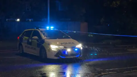 General view of the scene at night, with a police car and cordon in the rain
