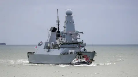 PA The HMS Dragon sailing through the sea. It is a large grey military ship with personnel stationed along it.