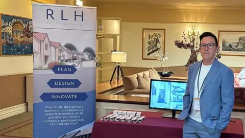 RLH Architectural Nick Cox standing in front of a stall showcasing his company - RLH Architectural. He is wearing a blue suit jacket and trousers and a white t-shirt. He has dark hair and glasses.