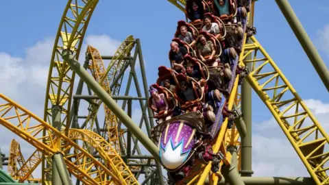 People in a purple rollercoaster race down a yellow track. There are loops and twists on the track behind them.