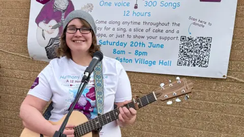 A woman wearing a T-shirt, glasses, and a grey beanie hat while holding a guitar and standing in front of a microphone. She is smiling and looking into a camera. 