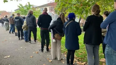 A long line of people waits single file on the side of a path, beside a patch of grass. They're waiting to get into a dentist's practice.