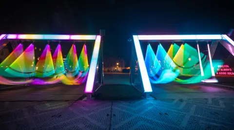Andre Pattenden The illuminated swing exhibit, taken using a low shutter speed to emphasise the movement of the piece. There are two separate swing sets side by side, lit up with blue, green, yellow and pink hues. 