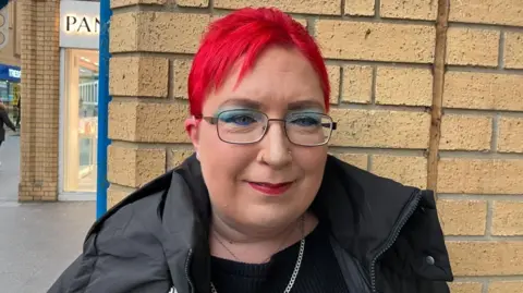 Vikki Irwin/BBC Jen Jones has bright red hair and is wearing a black coat and glasses. She is standing in front of a shop wall.
