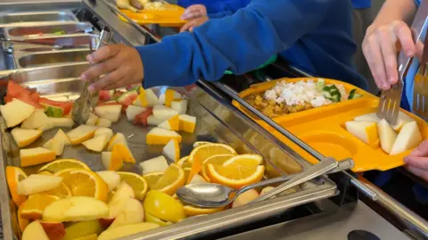BBC A large silver tray to the left is filled with a variety of fruit slices, including oranges and melon varieties. Two children's hands are holding utensils and placing items on yellow lunch trays to the right.