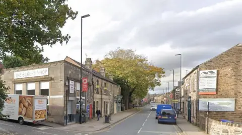 A picture of a street showing several shops and houses with vehicles parked on the road. 