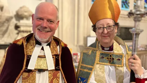 Jon Wright/BBC A dean and a bishop in full Anglian regalia smile at the camera 