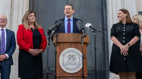 Welsh government Rhun ap Iorwerth, the first minister, standing at a lectern flanked by women and men, including Sioned Williams.