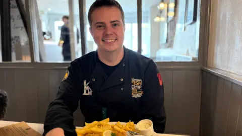 George sits in behind a plate of fish and chips, wearing his dark frying uniform