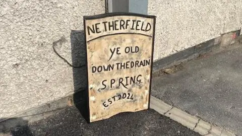 Dean Weatherbed/BBC A close up of the sign which reads "Netherfield. Ye Old Down The Drain Spring. Est 2024".