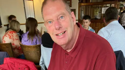 Tim Hull appears to be a bar. He smiles and wears a red shirt. His face seems complete than in other pictures