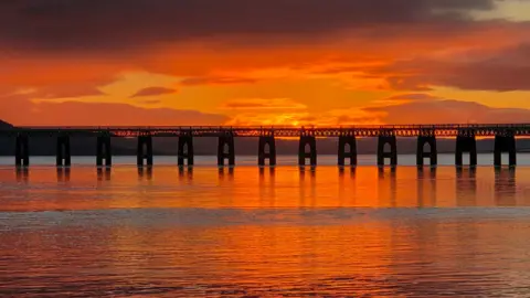 Shahzeb Saeed A long bridge stretches across calm water under a fiery orange sunset sky.
