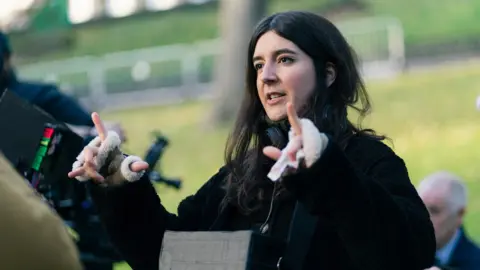 Conic Film Film maker Laura Carreira is standing in a park surrounded by film crew, seemingly giving directions during a film scene. Her hair is down and she wears a black thick jumper and gloves. 