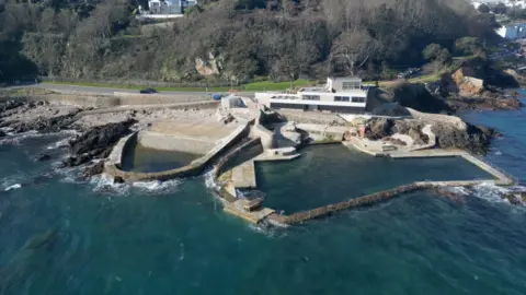 BBC Aerial view of La Vallette bathing pools in Guernsey, showing stone sea walls, a slipway and a rectangular tidal pool beside a coastal road and buildings above rocky shoreline.
