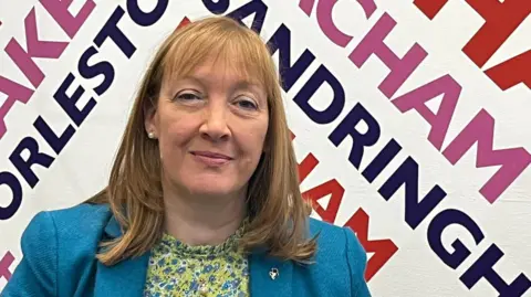 Nova Fairbank is looking at the camera, standing in front of a wall covered in names of local towns in a BBC studio. She has shoulder length brown hair and is wearing a patterned green blouse and a blue jacket.