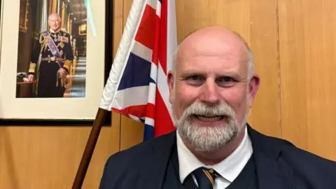 Tom Wootton with white hair and beard smiling at the camera and wearing a blue jacket, white shirt and blue tie. He is standing in front of a wooden wall hanging on which are a portrait of the King, and a union flag.