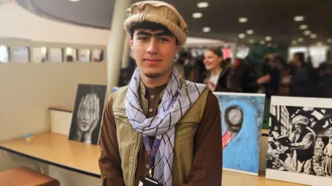 Eastern Education Group Omran is wearing a brown hat, a blue and white striped scarf, a beige gilet and a brown shirt. His college badge hangs around his neck on a lanyard. He is in a large atrium with paintings behind him, representing different cultures