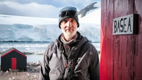 UKAHT/Pete Watson Carpenter Jo Oliver, a middle aged man in cold weather gear, standing in the Antarctic beside a wooden wall with a sign saying 'Base A' and a wooden hut in the background. In the distance are snow covered mountains and a glacier.