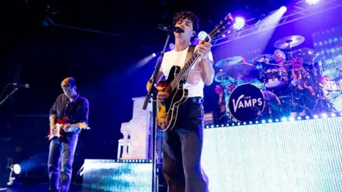 Getty Images Two members of The Vamps holding guitars and performing on stage with microphones and a drum band in the background