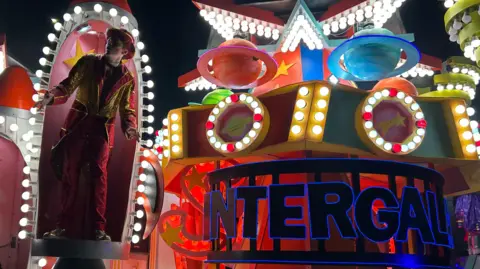 A carnival cart. There is a male performer standing under an arch on the cart, next to a display that features two small planets.