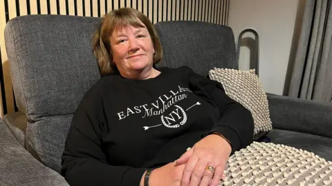 A woman wearing a black jumper. She has her hands on her lap and a brown bob.