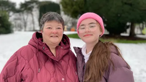 Isla and Kimberly standing side by side. Isla has very long brown hair which is tied back and is wearing a pink hat, silver glasses, purple puffer coat and jumper. Kimberly has short dark hair and is wearing a red coat.