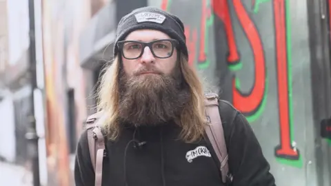 Tom is looking towards the camera. He is wearing a black hoodie, black beanie and black glasses. He is also wearing a light pinkish backpack. 