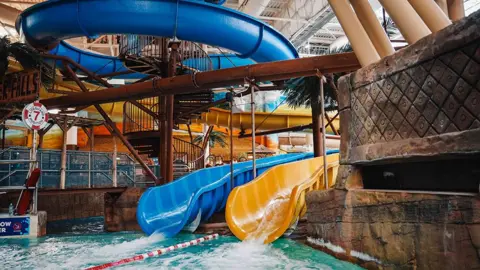 A blue and a yellow water slide next to each other opening into a pool at the Sandcastle. There is a big loop of the blue slide at the top and the yellow one is seen further in the distance.
