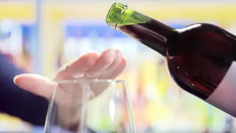 A person puts their hand over the top of a wine glass as wine is to be poured from a bottle.