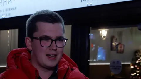 Jack Taylor-Walker in Stafford High Street at night, with a shop window behind him, wearing a red jacket and glasses.