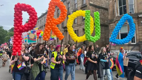 Pride march in Glasgow street. Marchers are carrying colourful letters made up of  red, orange, yellow, green and blue balloons that spell out the word PROUD