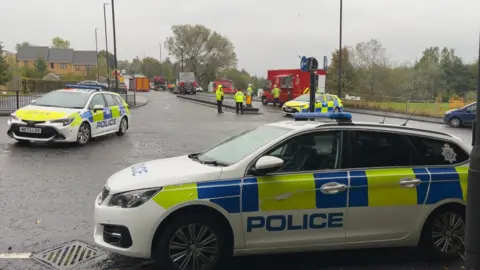 Police cars close the road. Fire engines can be seen in the background. It is day time and cloudy.