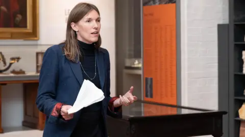 Faye Sayer A woman with long brown hair, a blue jacket, necklace, and black top, stands in a room and speaks while holding a piece of paper in her right hand.